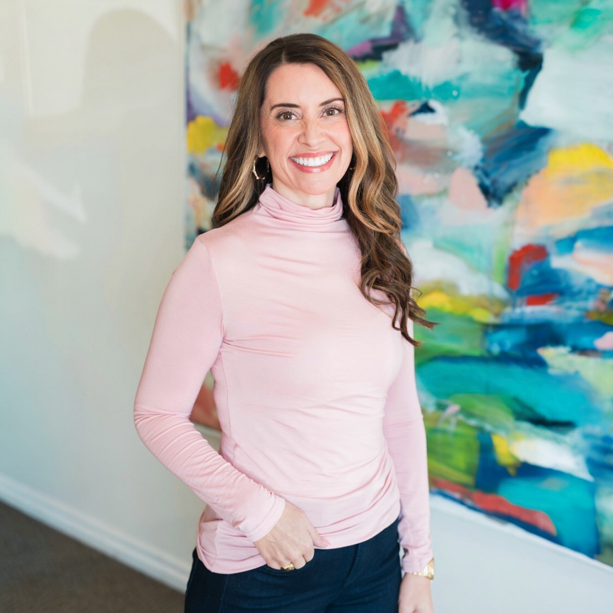 Woman wearing a pink turtleneck sweater standing in front of a colorful abstract painting.