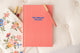 Kind Birdie The Aligned Journal, a coral pink guided journal with blue text, next to a floral embroidered pouch and a coral pen.
