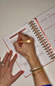 Kind Birdie Upward Planner: A woman's hand with coral nails writes in a 90-day Christian planner with gold coil binding.