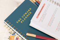 Kind Birdie Upward Planner Annual Bundle, a teal planner with gold lettering and a spiral binding, next to an open planner with daily focus prompts.