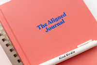 The Aligned Journal, a coral-colored guided journal with blue text, and a white Kind Birdie pen.