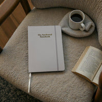 Kind Birdie The Anchored Notebook, a light grey blank journal with gold lettering, sits on a textured chair next to a cup of coffee and an open book.